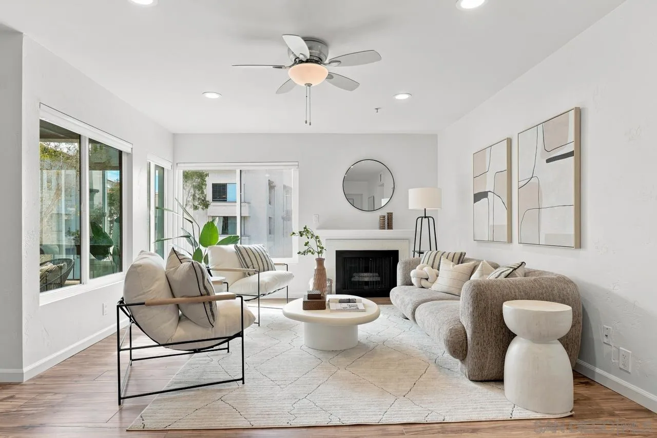 Staged living room with a fireplace, large windows, neutral seating, and a ceiling fan.