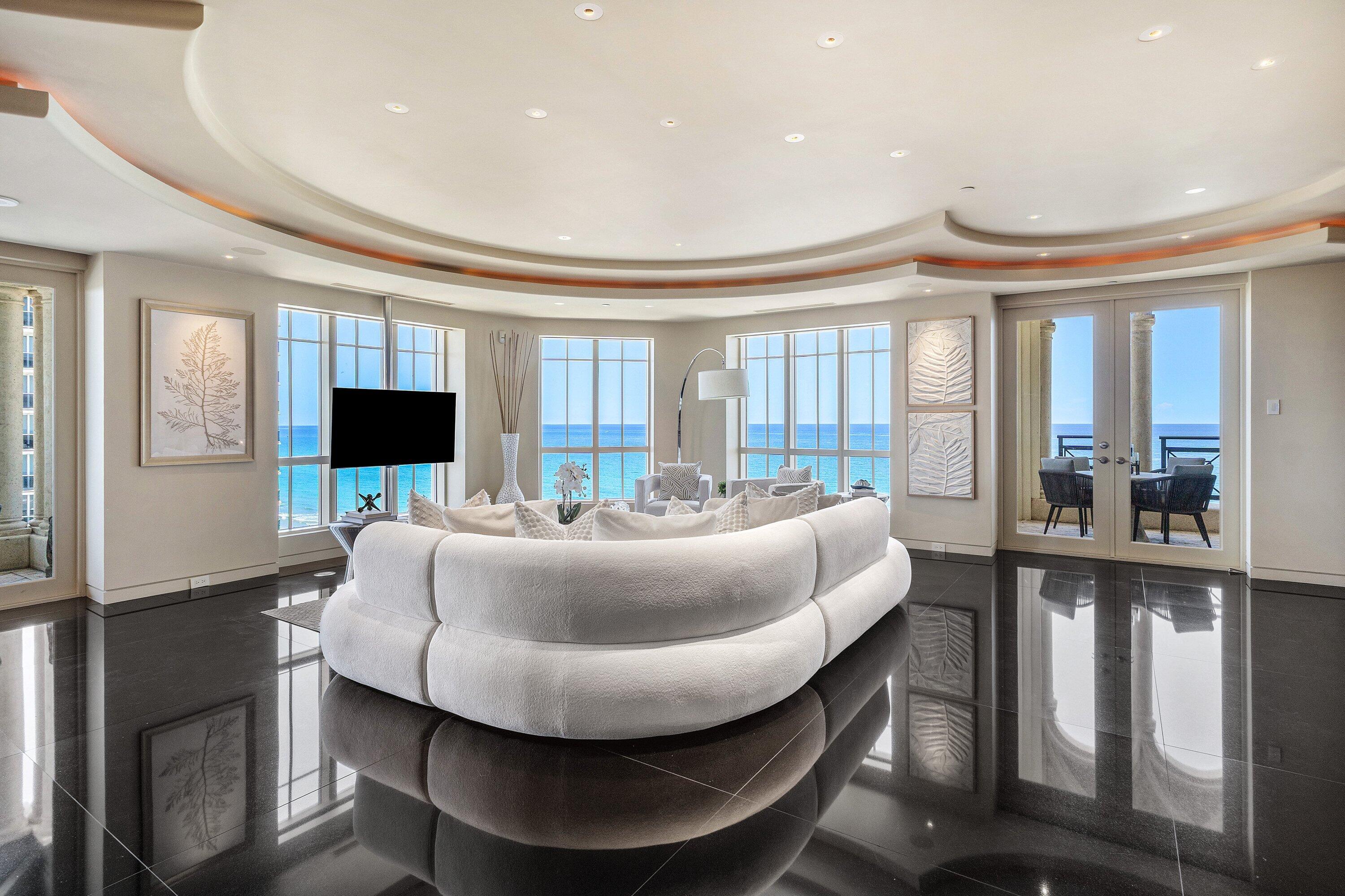Oceanfront living room with a curved white sectional, glossy floors, and balcony doors overlooking the water.