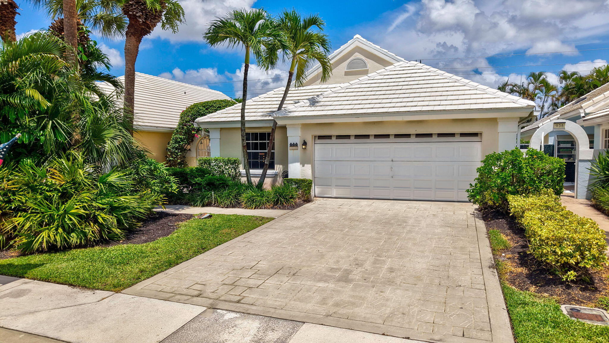 Cream stucco home with a tiled roof, double garage, paved driveway, and tropical landscaping.