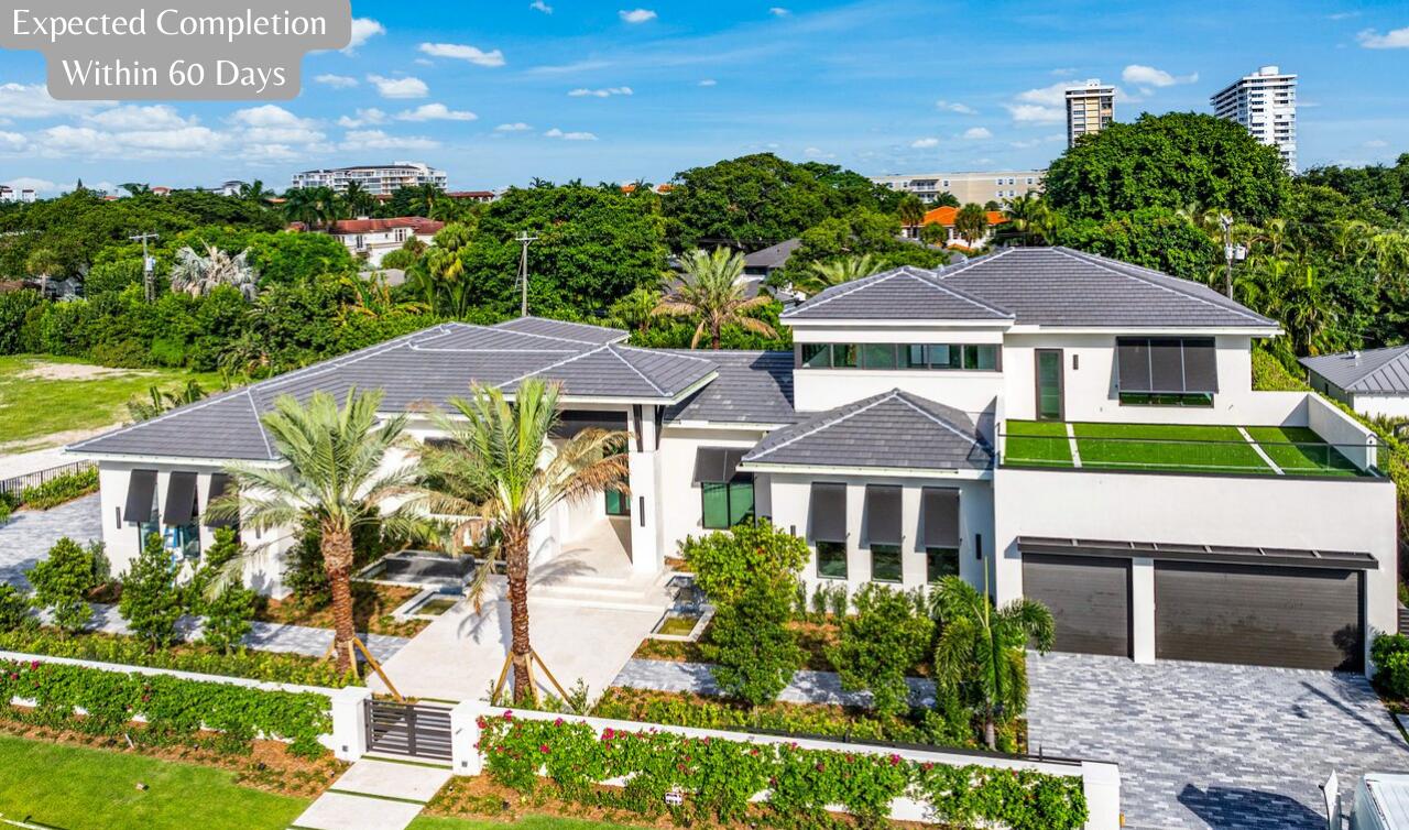 Modern white home with gray tile roofs, palm-lined grounds, and a gated driveway.