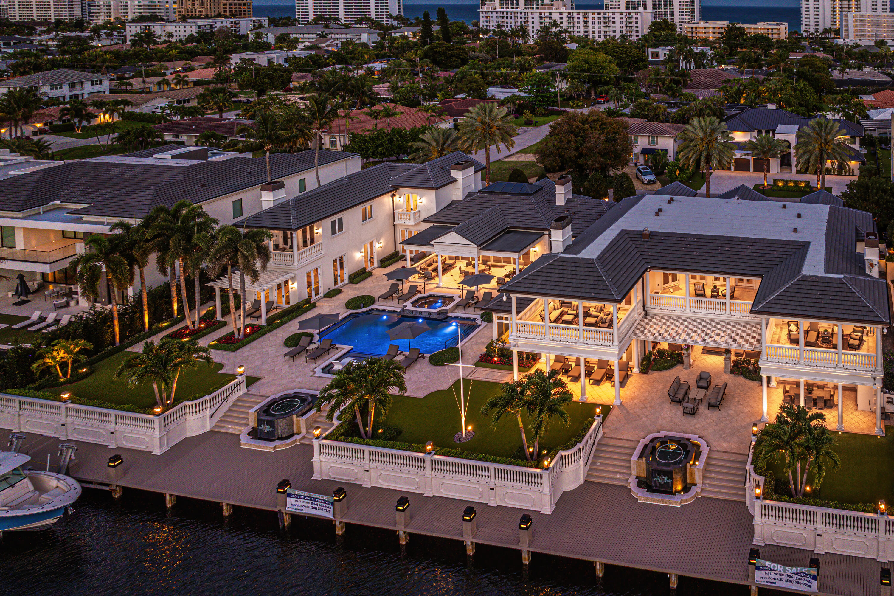 Waterfront home with illuminated patios, a swimming pool, palm trees, and a long dock at dusk.