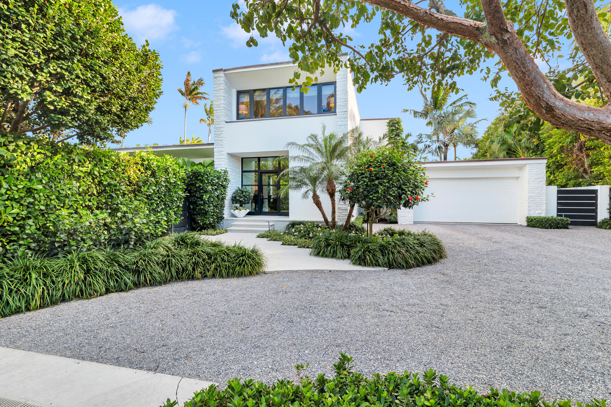 Modern white two-story home with a gravel driveway, attached garage, and tropical landscaping.