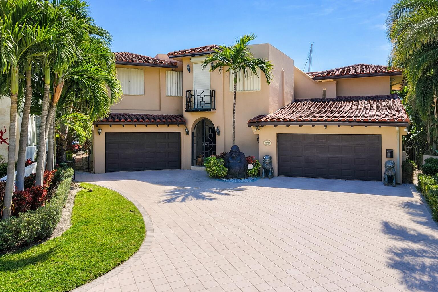 Beige stucco home with two garages, red tile roofs, palm trees, and a broad paved driveway.