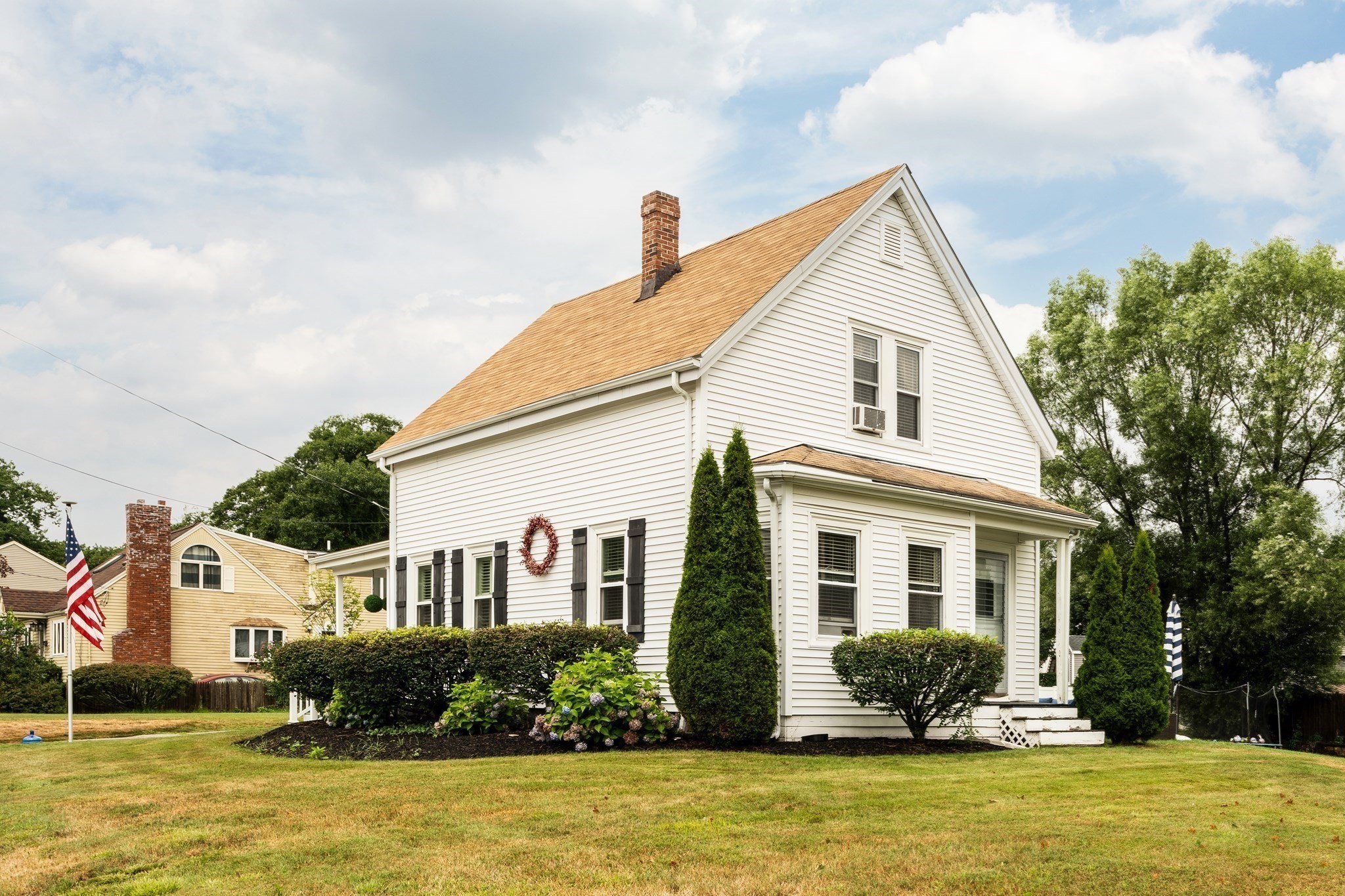 White clapboard house with a steep gabled roof, brick chimney, and landscaped lawn.