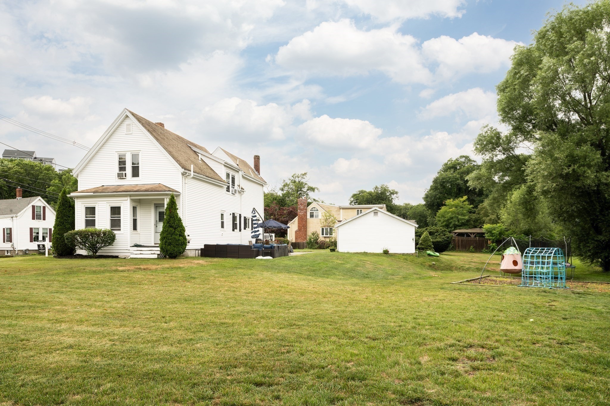 White two-story house behind a broad lawn, with patio seating, a detached outbuilding, and a children's playset.