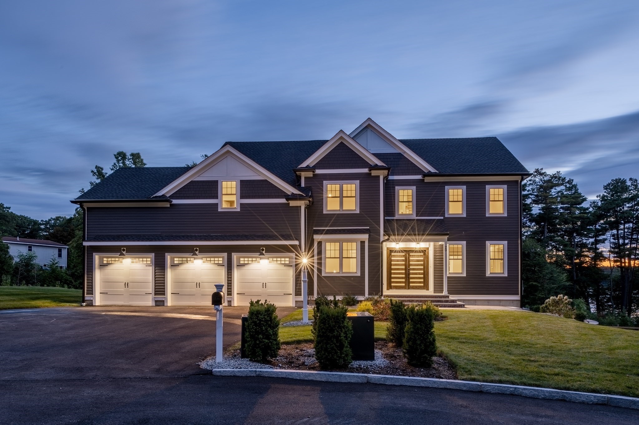 Two-story dark-sided house with three-car garage and illuminated windows at dusk.