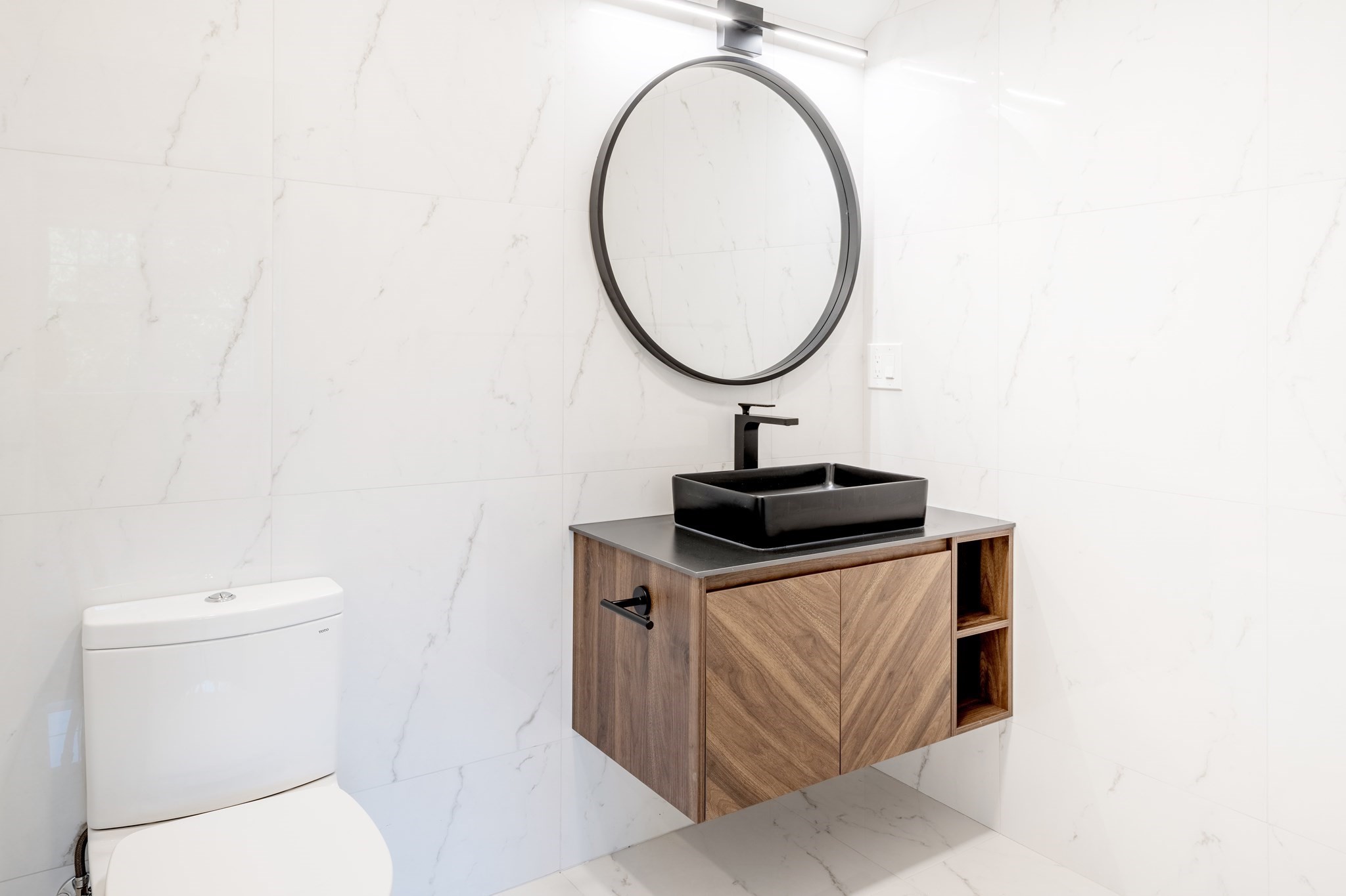 Modern bathroom with a floating wood vanity, black vessel sink, round mirror, and white marble-look tile.