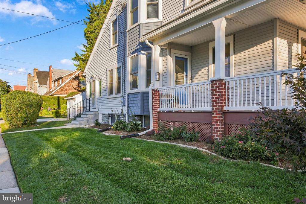 Two-story gray-sided house with a covered porch, white railings, brick supports, and a green front lawn.