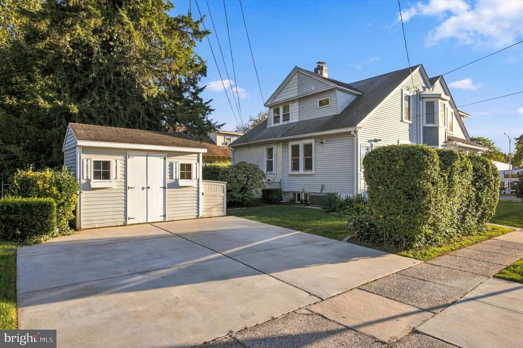 White gabled house with a detached shed, wide concrete driveway, and trimmed hedges.