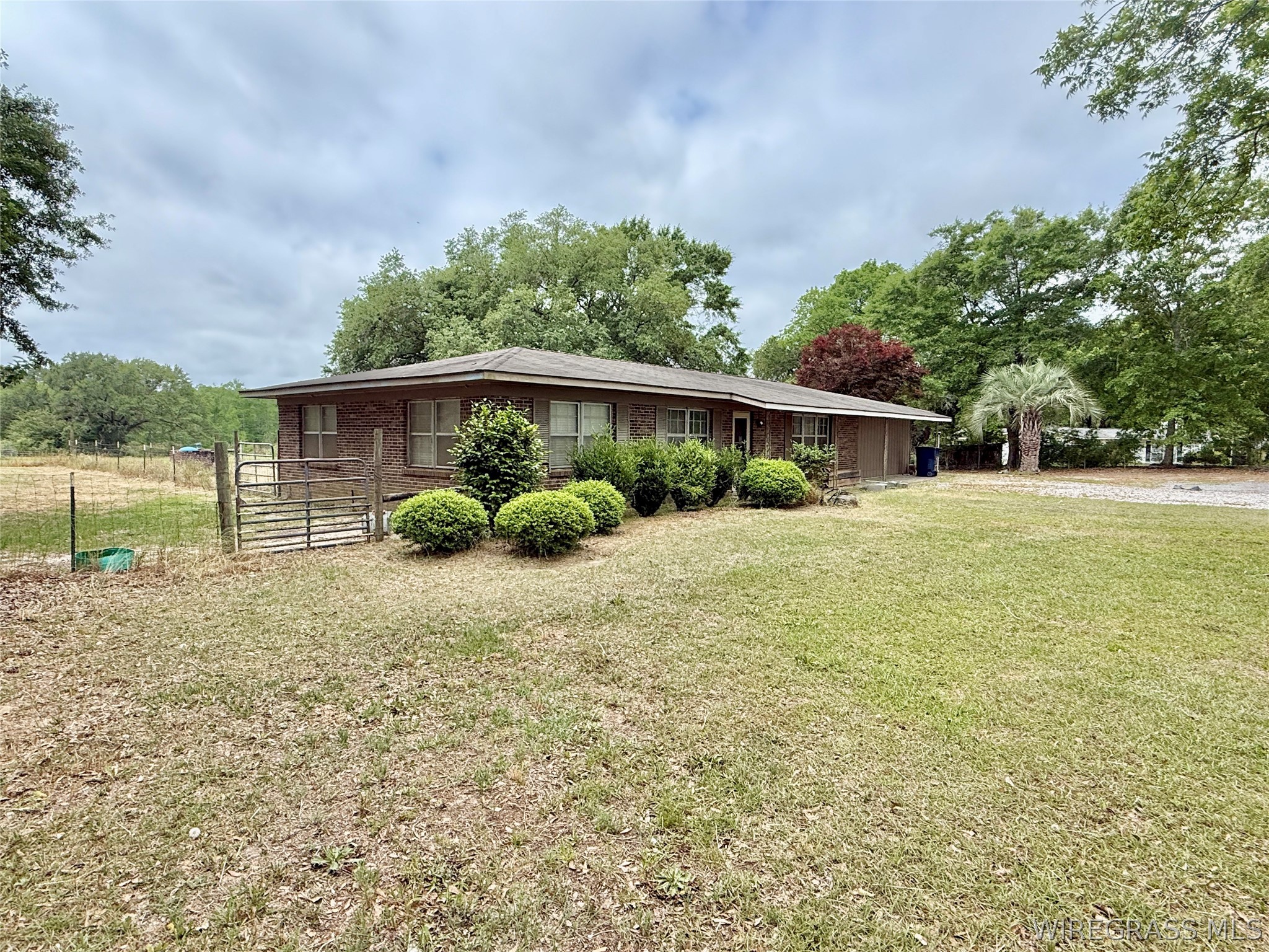 Single-story brick house with front shrubs, a broad lawn, metal gate, and mature trees.