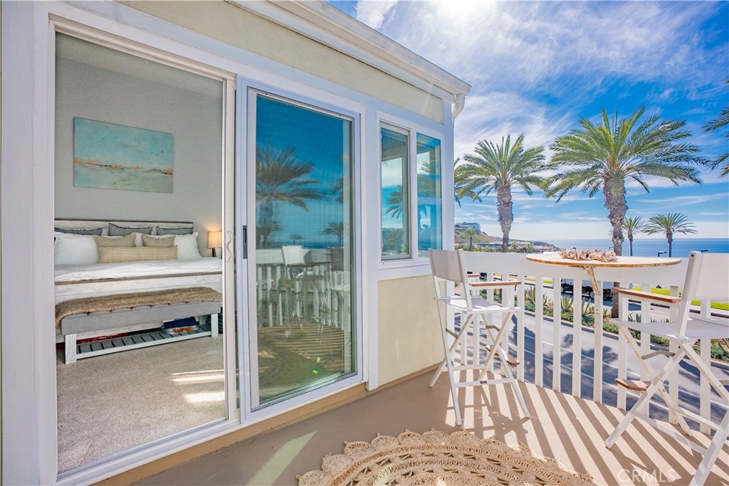 Condo balcony with white dining furniture overlooking palm trees and the ocean, beside an open bedroom.