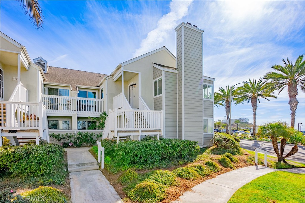 Residential building with white-railed balconies, landscaped shrubs, and palm trees beside the sidewalk.
