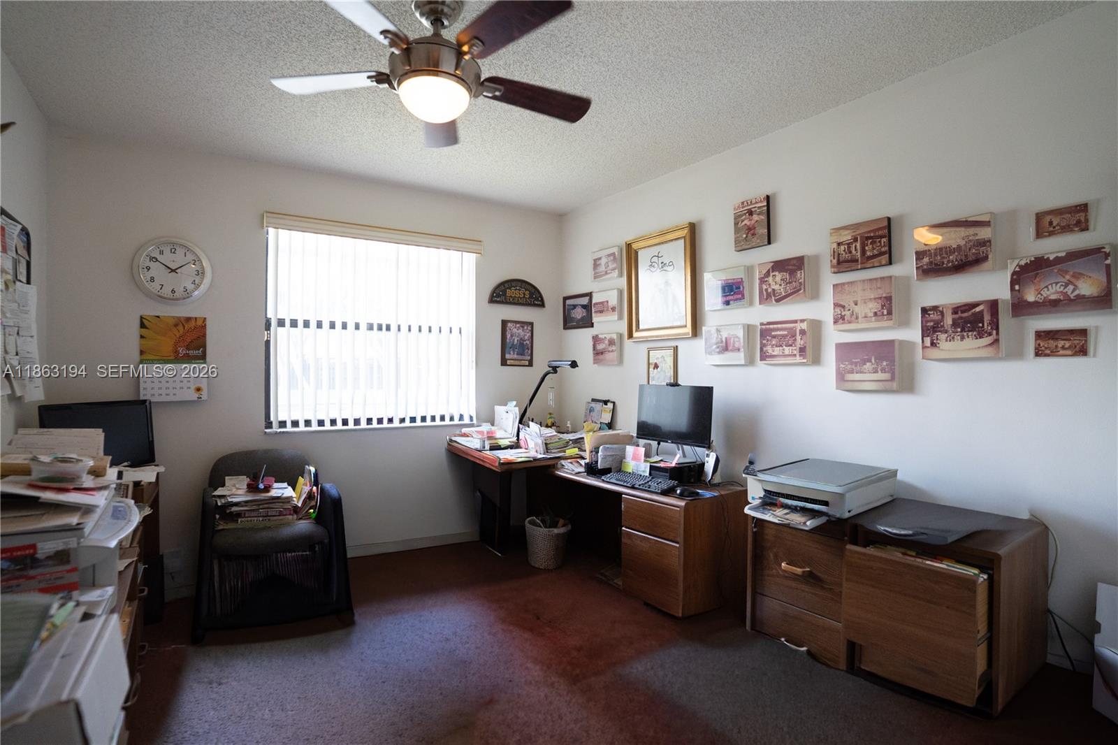 Cluttered home office with wooden desks, computer equipment, and framed pictures covering one wall.