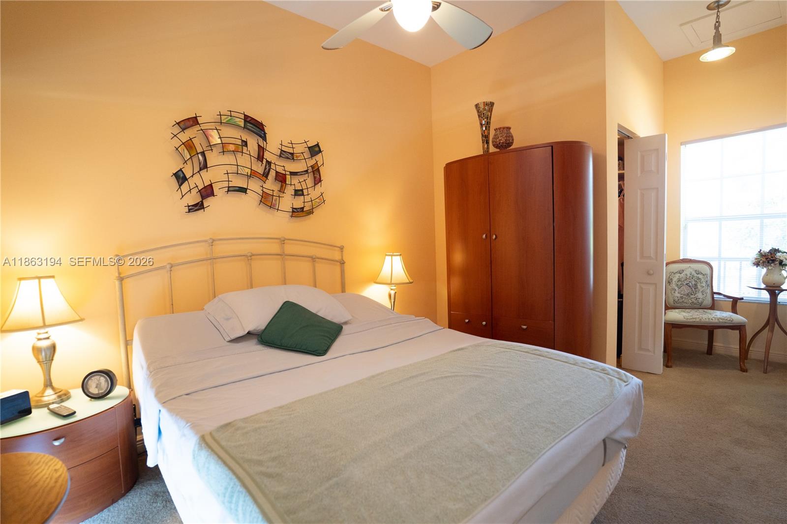 Furnished bedroom with a large bed, wooden wardrobe, and a chair beside the window.