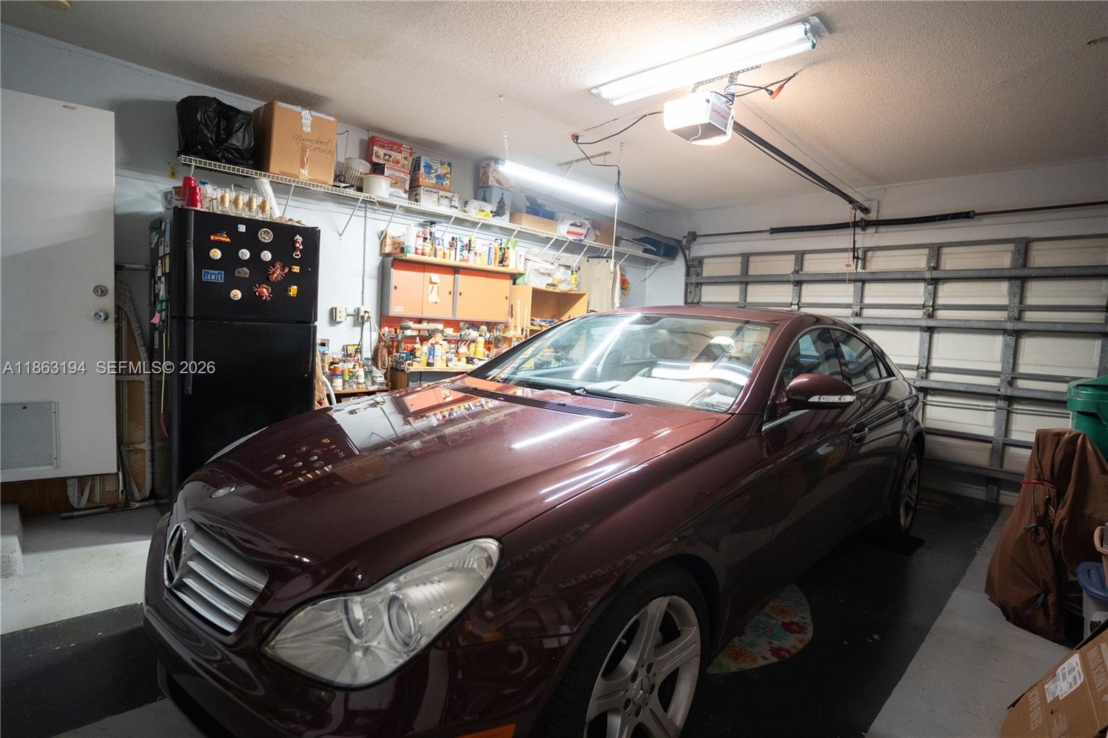 Garage with a burgundy sedan, storage shelves, refrigerator, and closed sectional door.