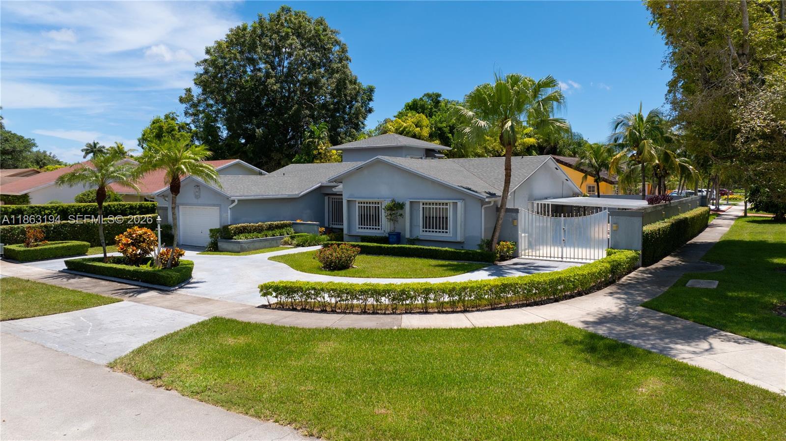 Single-story blue-gray house with a gated driveway, manicured lawn, hedges, and palm trees.