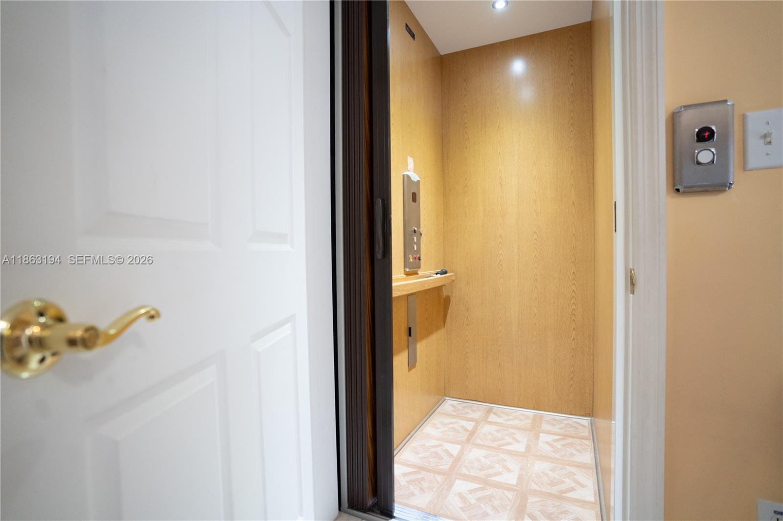 Open residential elevator with warm wood paneling, patterned floor, and white entry door.