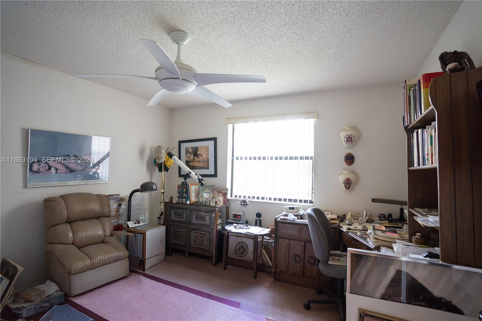 Furnished home office with cluttered desks, wooden bookcases, a beige recliner, and a ceiling fan.
