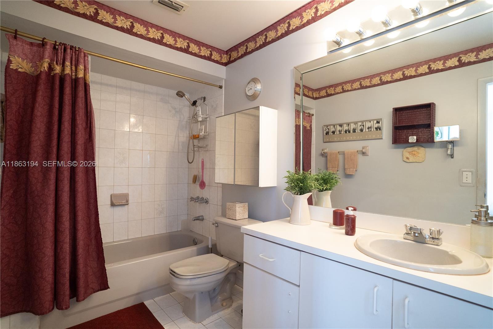 Bathroom with a tiled tub and shower, burgundy curtain, and long white vanity beneath a large mirror.