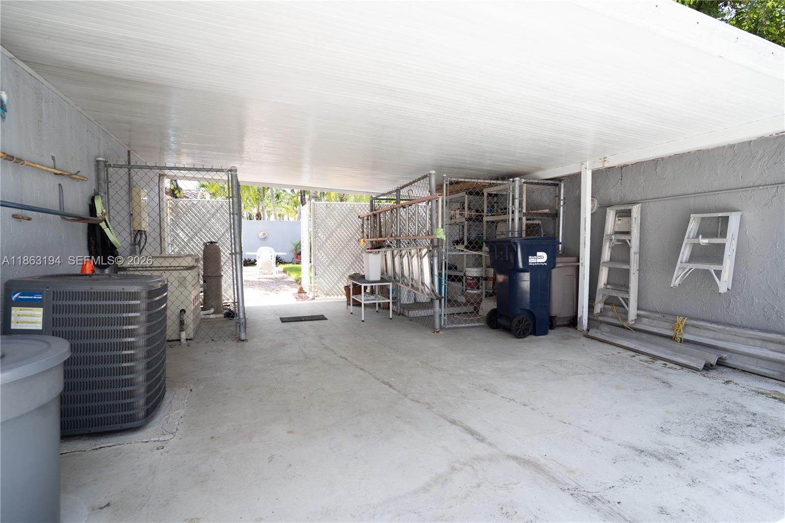 Covered carport with chain-link storage enclosures, air-conditioning equipment, and an open gate to the yard.