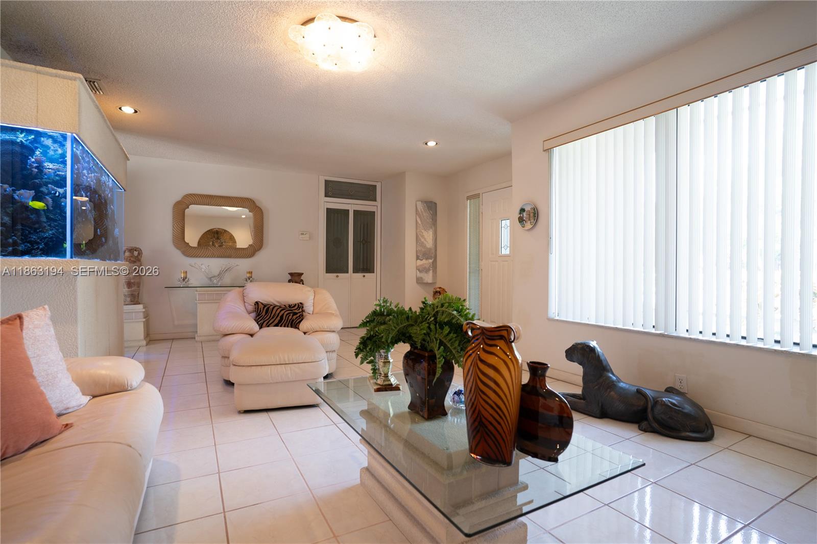 Living room with cream leather seating, a glass coffee table, and a large aquarium beside vertical blinds.