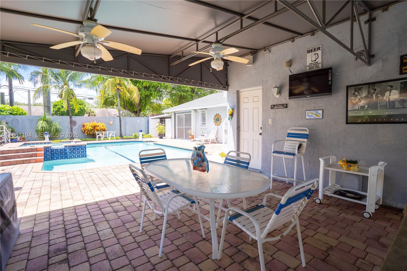 Covered poolside patio with ceiling fans, brick pavers, and a dining table beside the swimming pool.