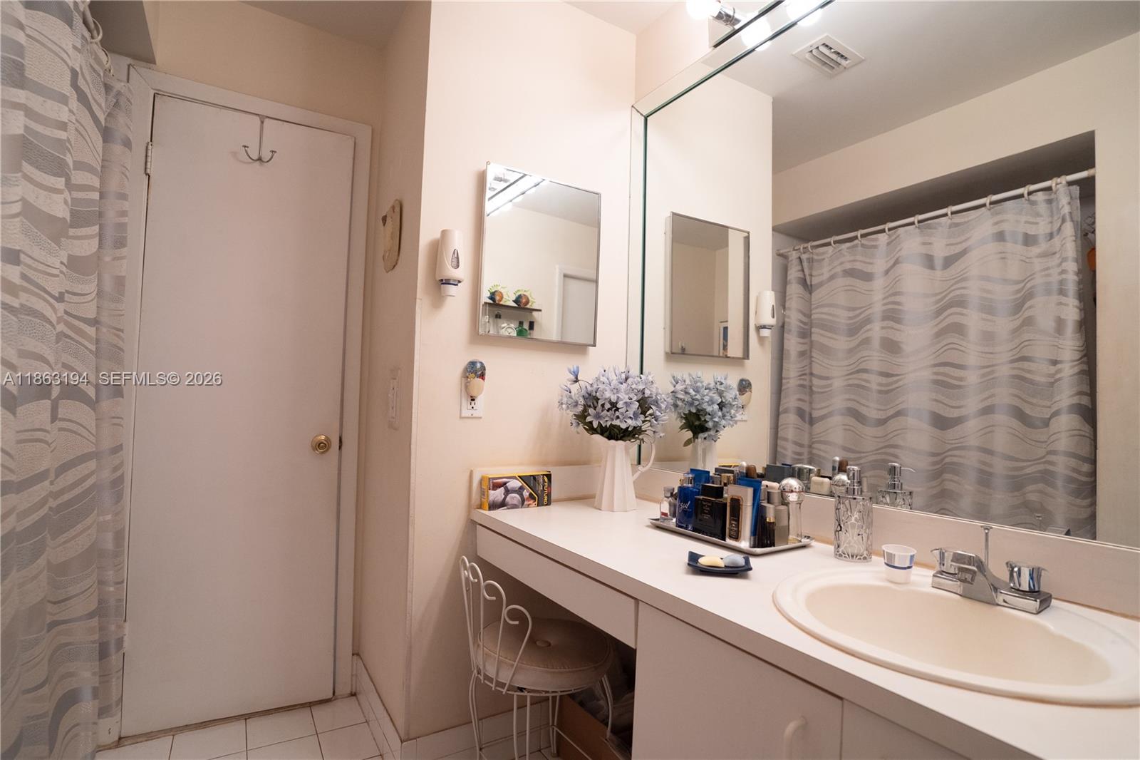 Bathroom vanity with oval sink, large mirror, flowers, and gray patterned shower curtain.
