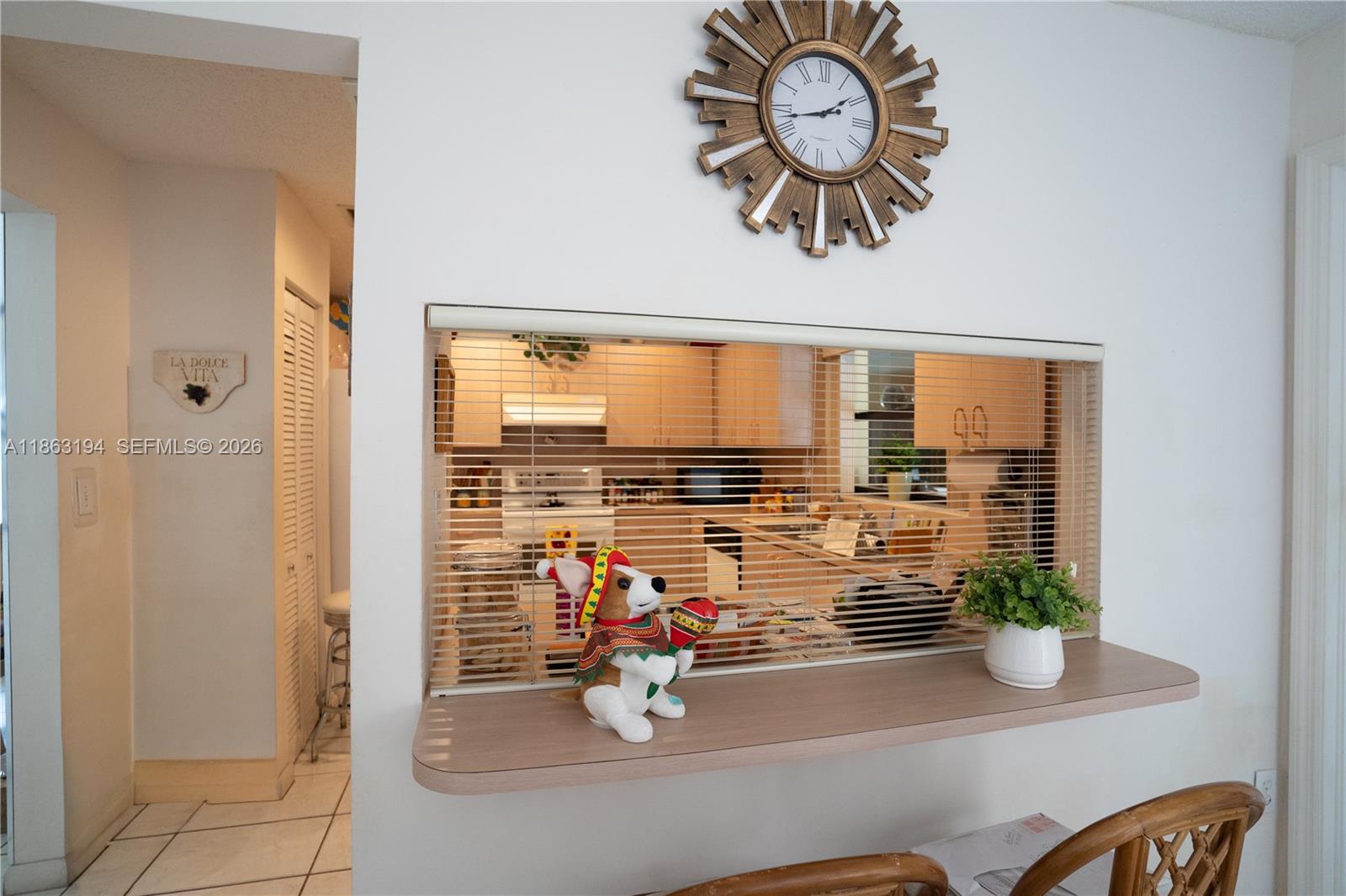 Kitchen viewed through a pass-through, with blinds, a sunburst clock, and decorative items on the ledge.