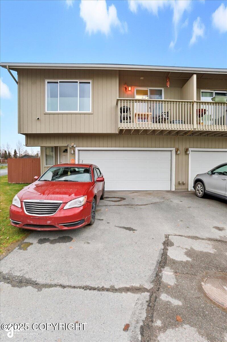 Beige multi-unit building with a second-floor balcony, two garage doors, and cars in the driveway.
