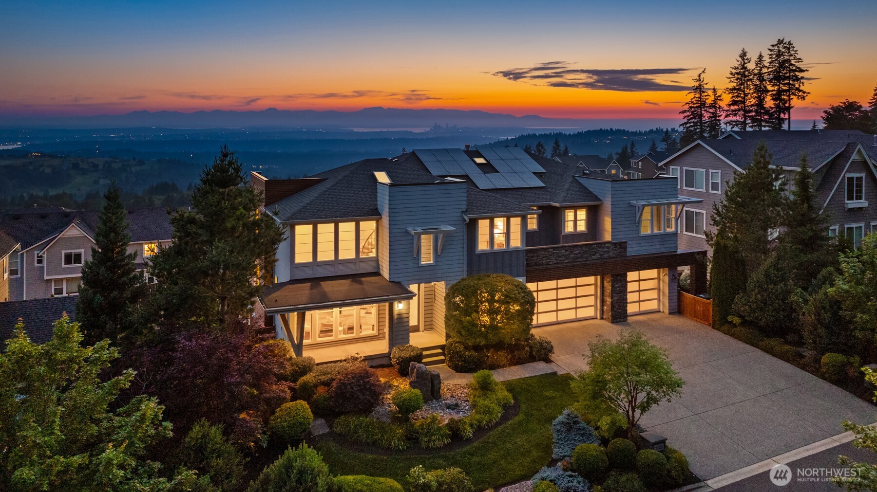 Modern home with solar panels overlooking water, a distant city skyline, and mountains at sunset.