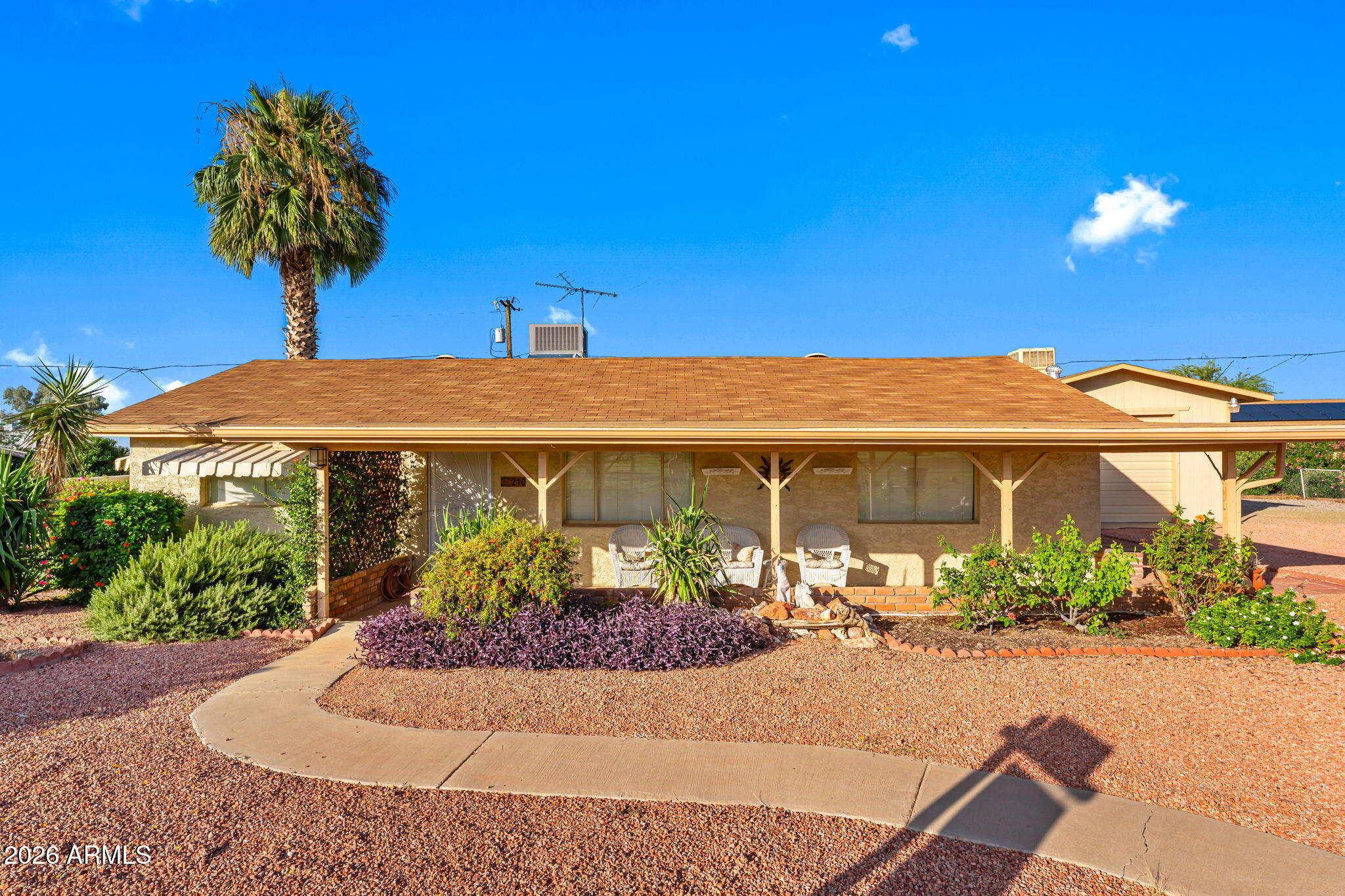 Single-story tan home with a covered porch, gravel yard, palm tree, and curved walkway.