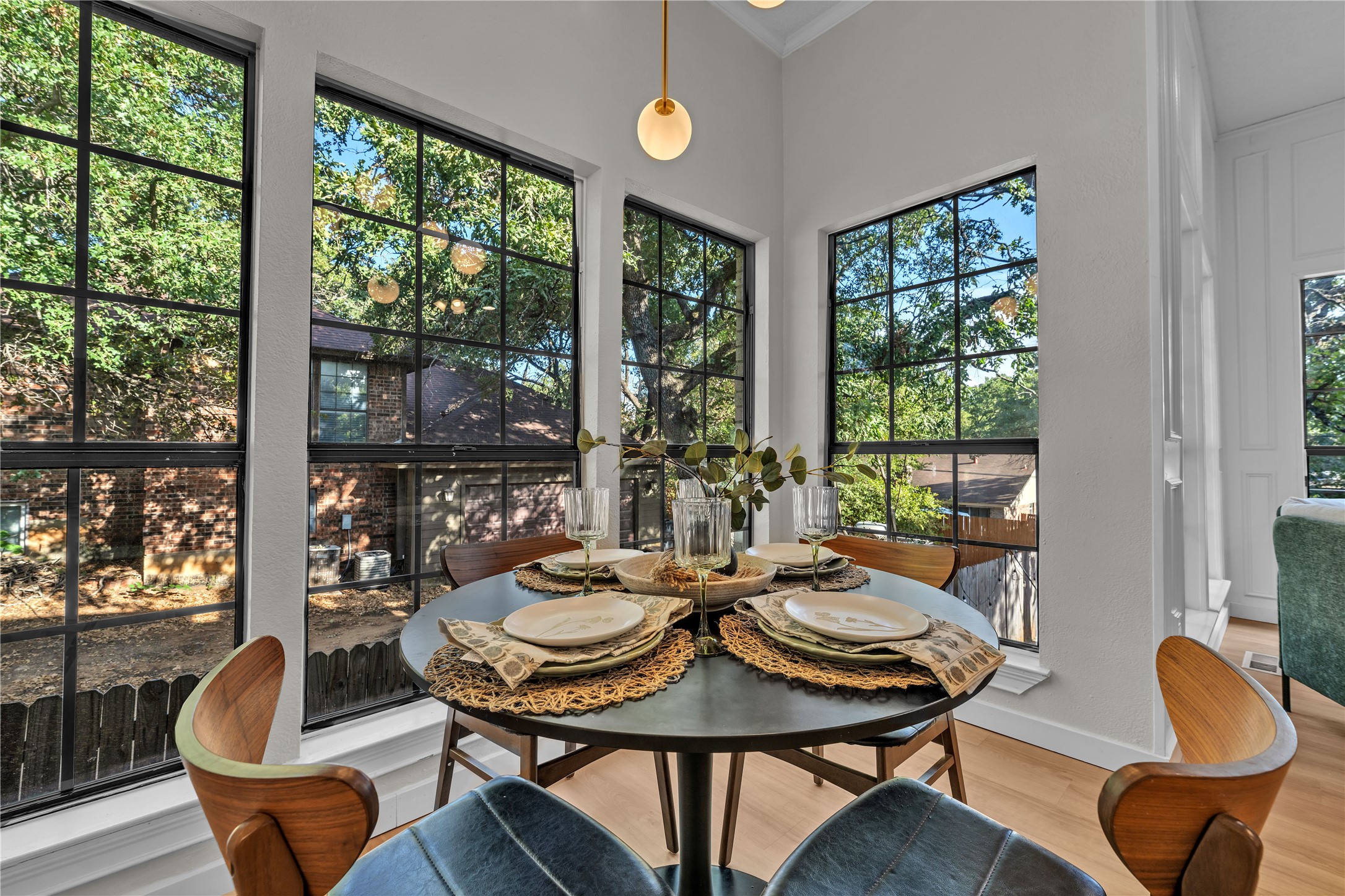 Dining area with a set round table and tall black-grid windows overlooking leafy trees.