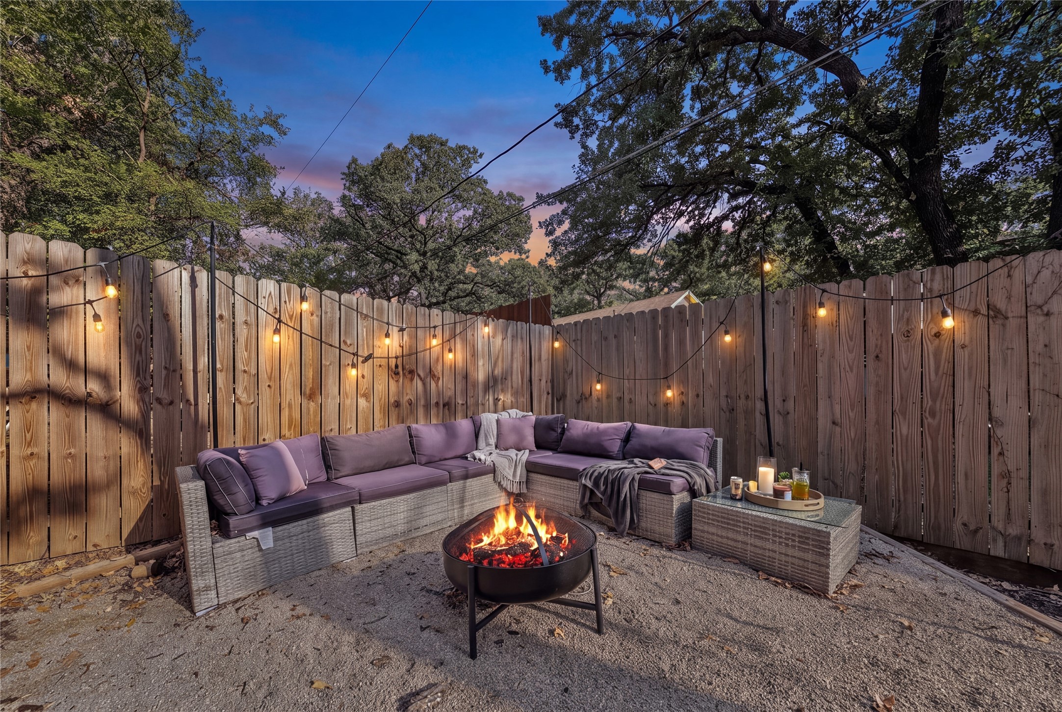 Backyard patio with a purple-cushioned sectional, glowing fire pit, and string lights at dusk.