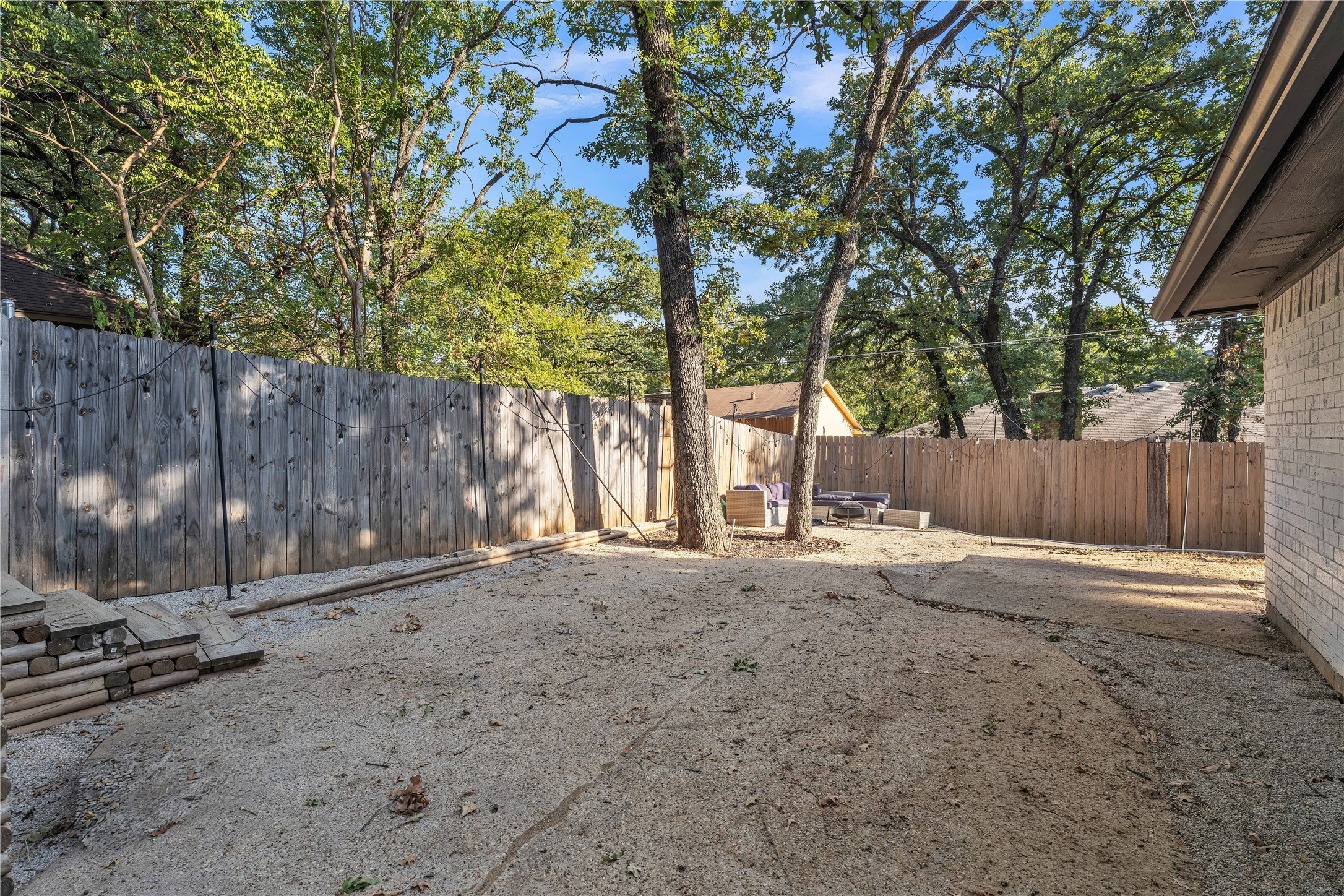 Fenced backyard with mature trees, gravel ground, string lights, and a small outdoor seating area.