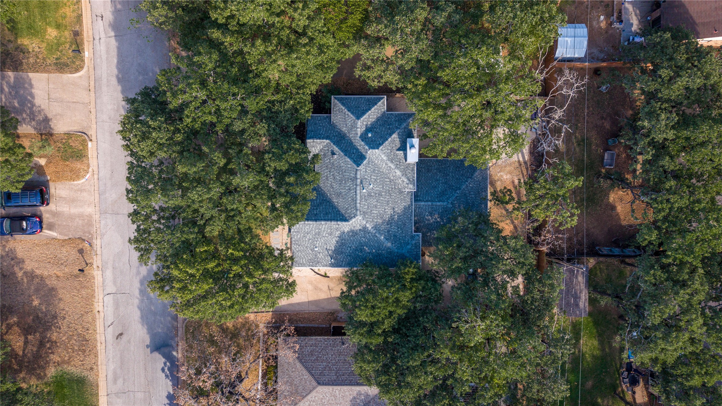 Aerial view of a gray-shingle-roofed house surrounded by mature trees beside a residential street.