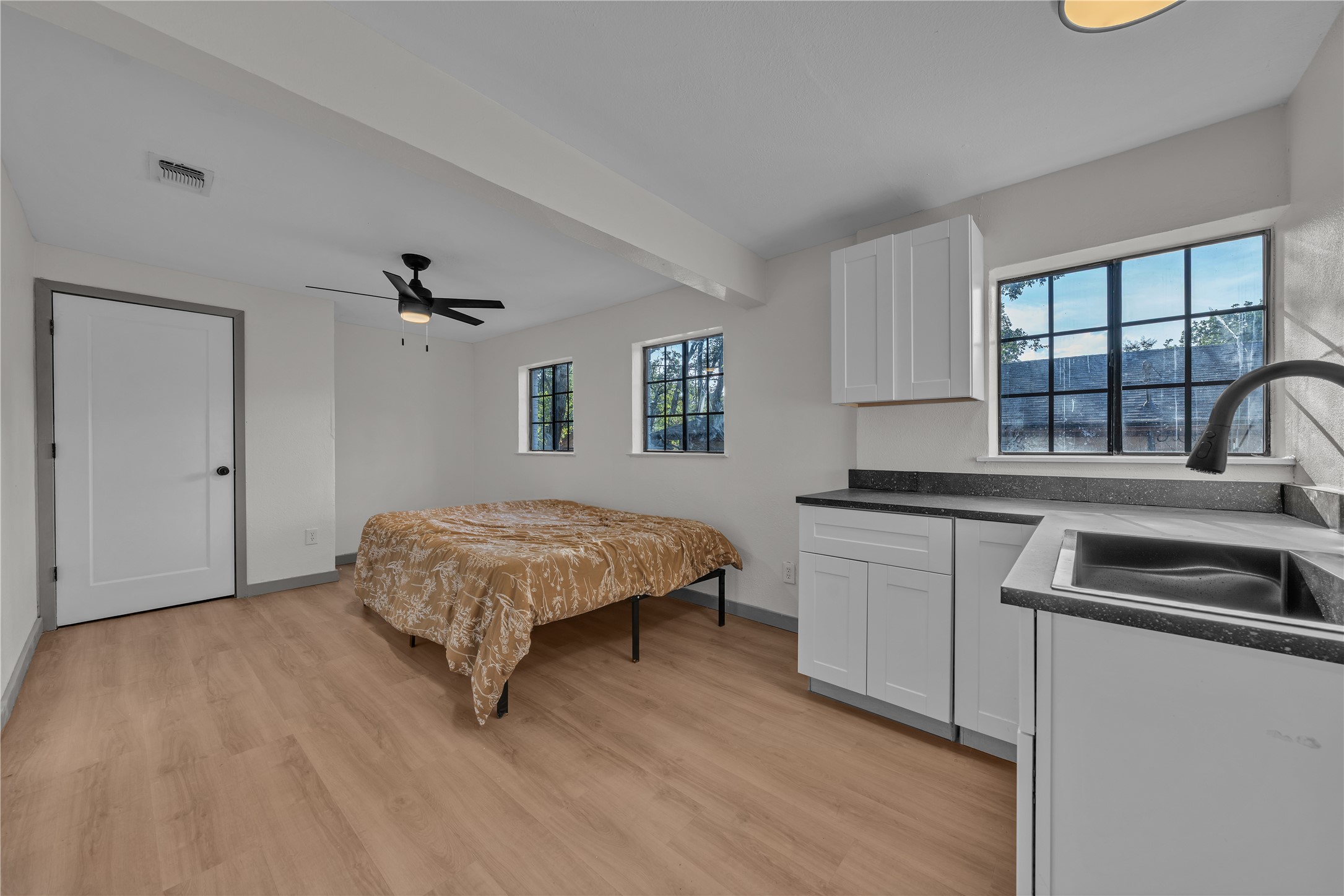 Bedroom with a bed beside a compact kitchenette, white cabinetry, dark counters, and black-framed windows.