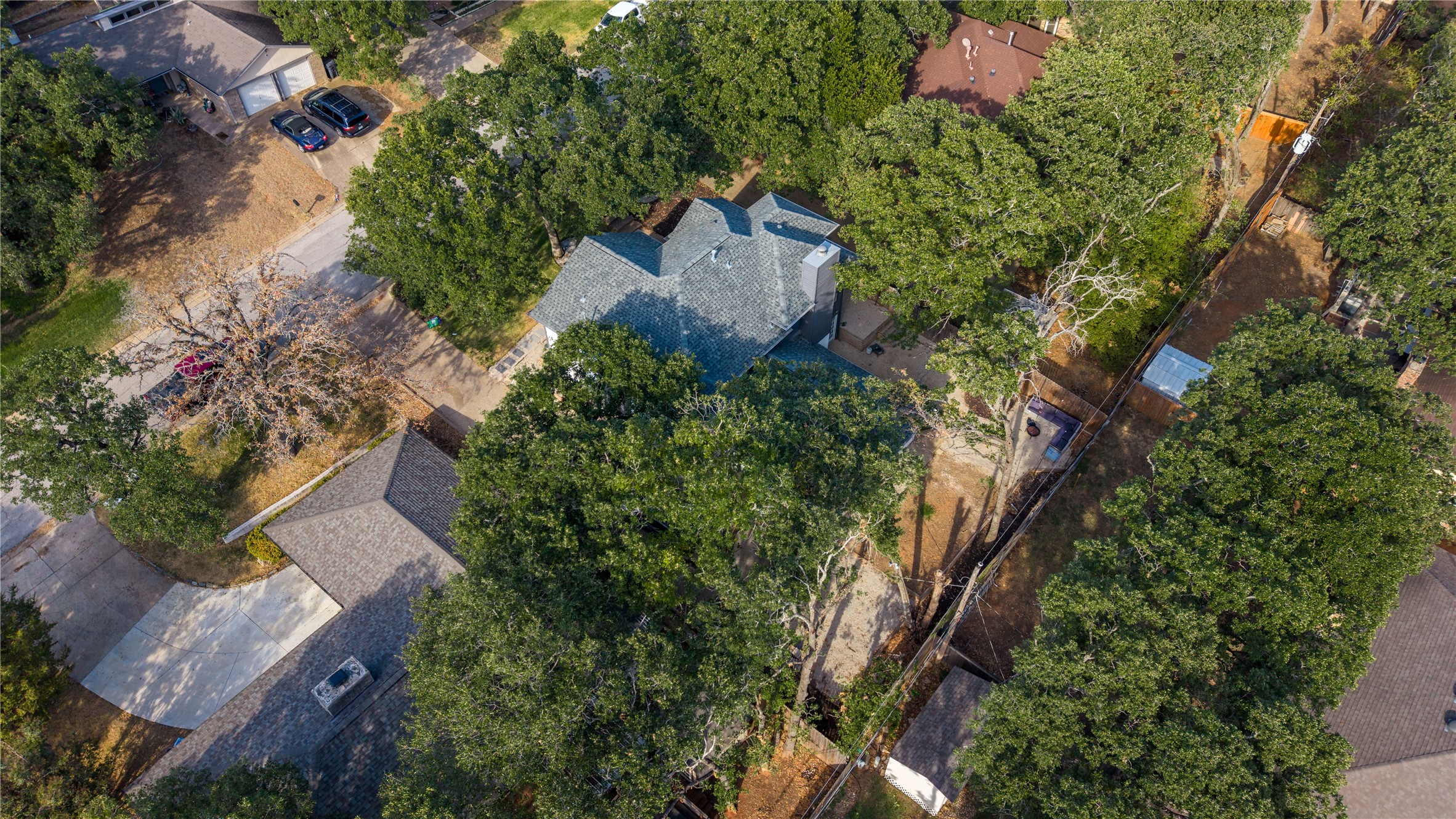 Aerial view of a gray-shingle-roofed home surrounded by mature trees and neighboring houses.