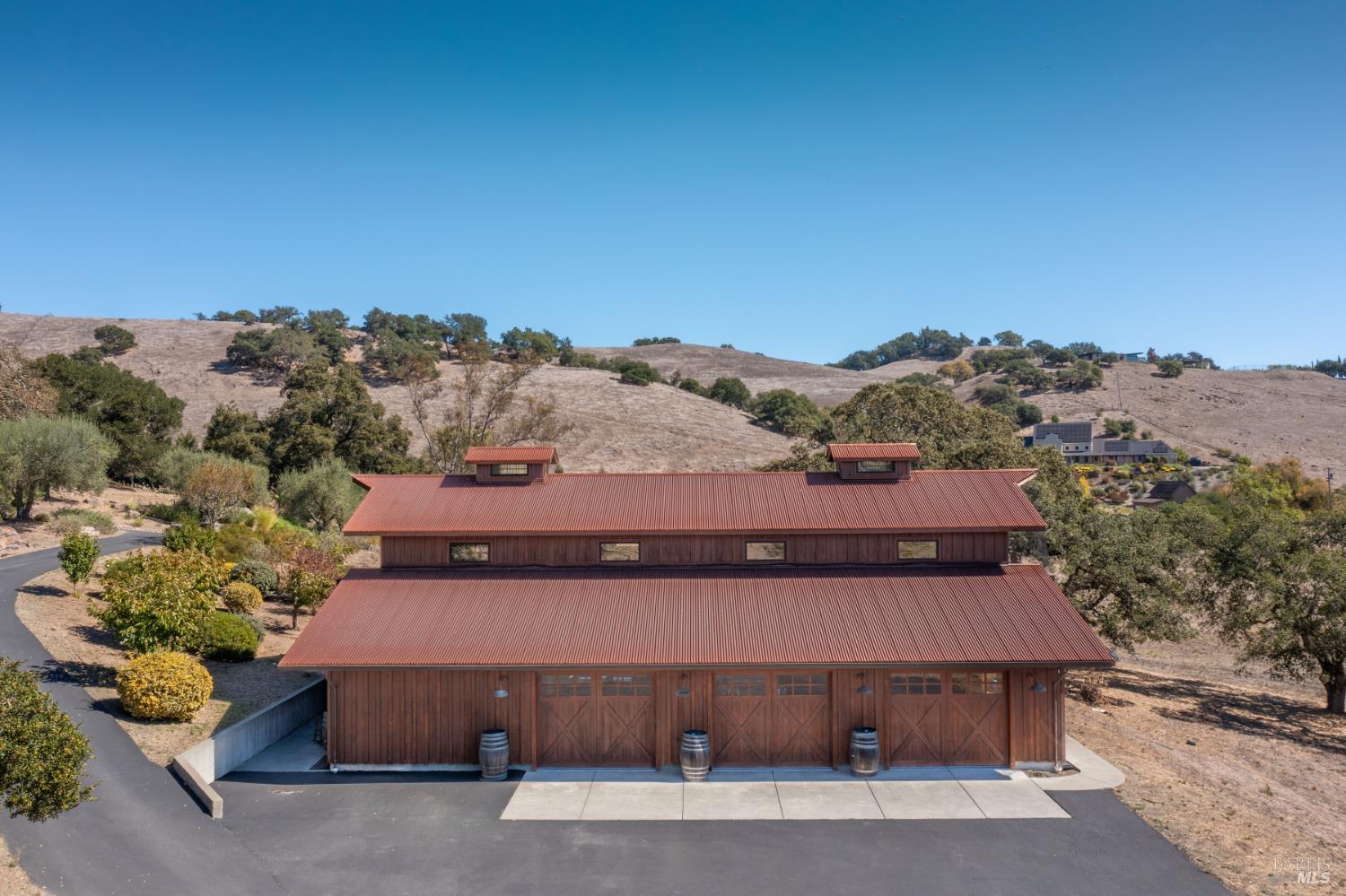 Barn-style property with red metal roofs, multiple garage doors, and dry rolling hills behind it.