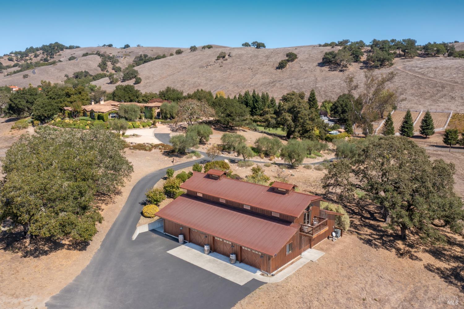 Aerial view of a brown-roofed building and winding driveway amid landscaped grounds and dry rolling hills.