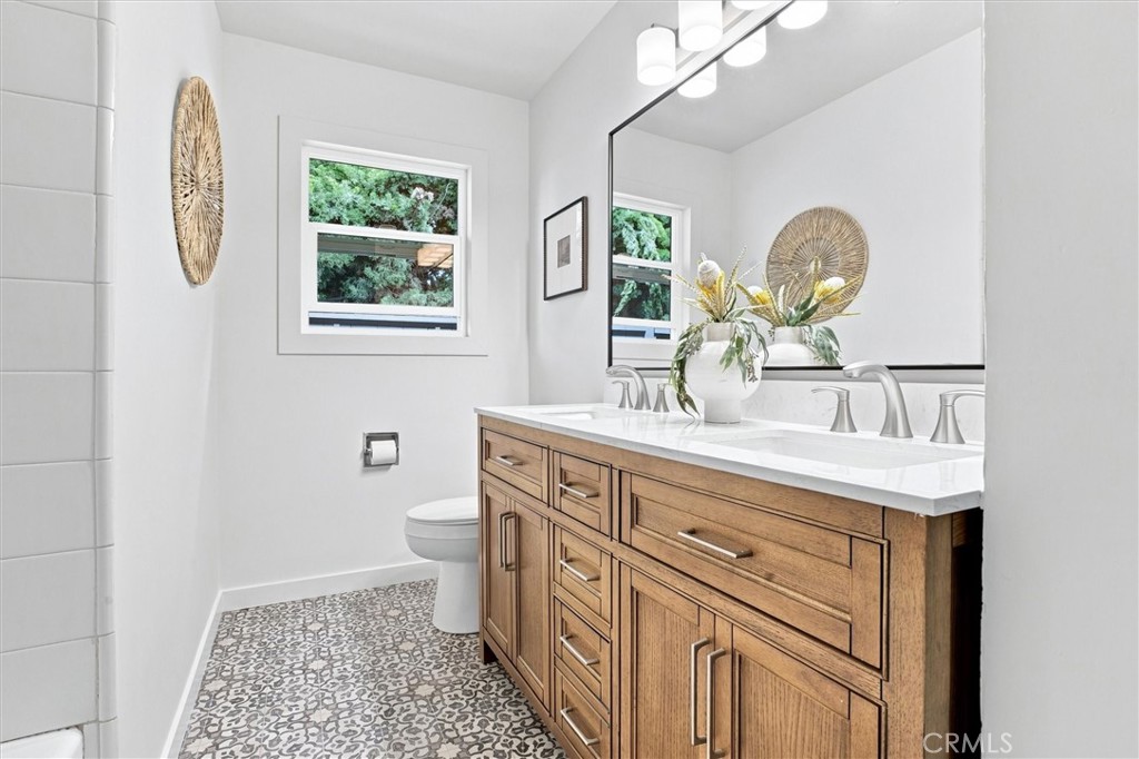 Bathroom with a double-sink wood vanity, patterned tile floor, and window overlooking greenery.