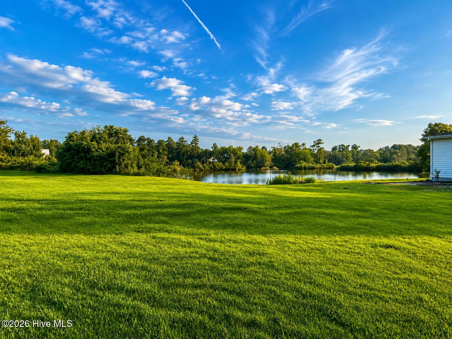Green lawn beside a pond, with a tree-lined opposite bank and part of a building at right.