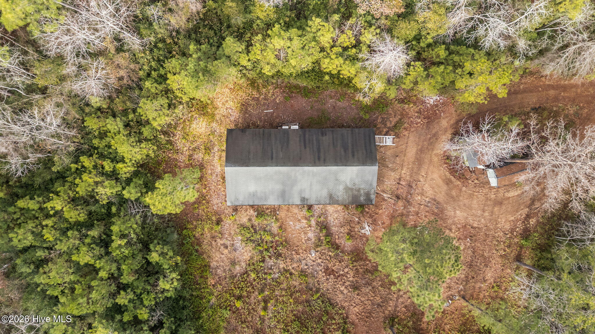 Aerial view of a dark-roofed house amid a wooded lot with a broad cleared area.