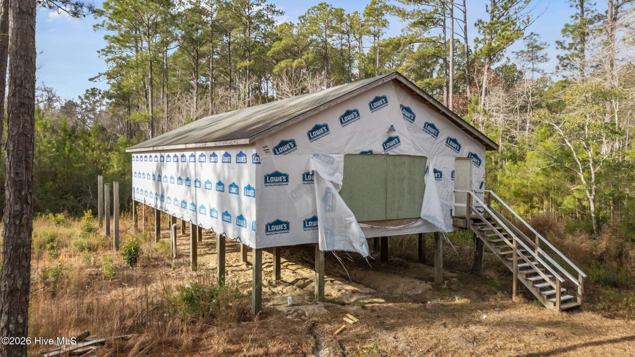 Elevated house under construction in a wooded lot, wrapped in building material with stairs at the side.