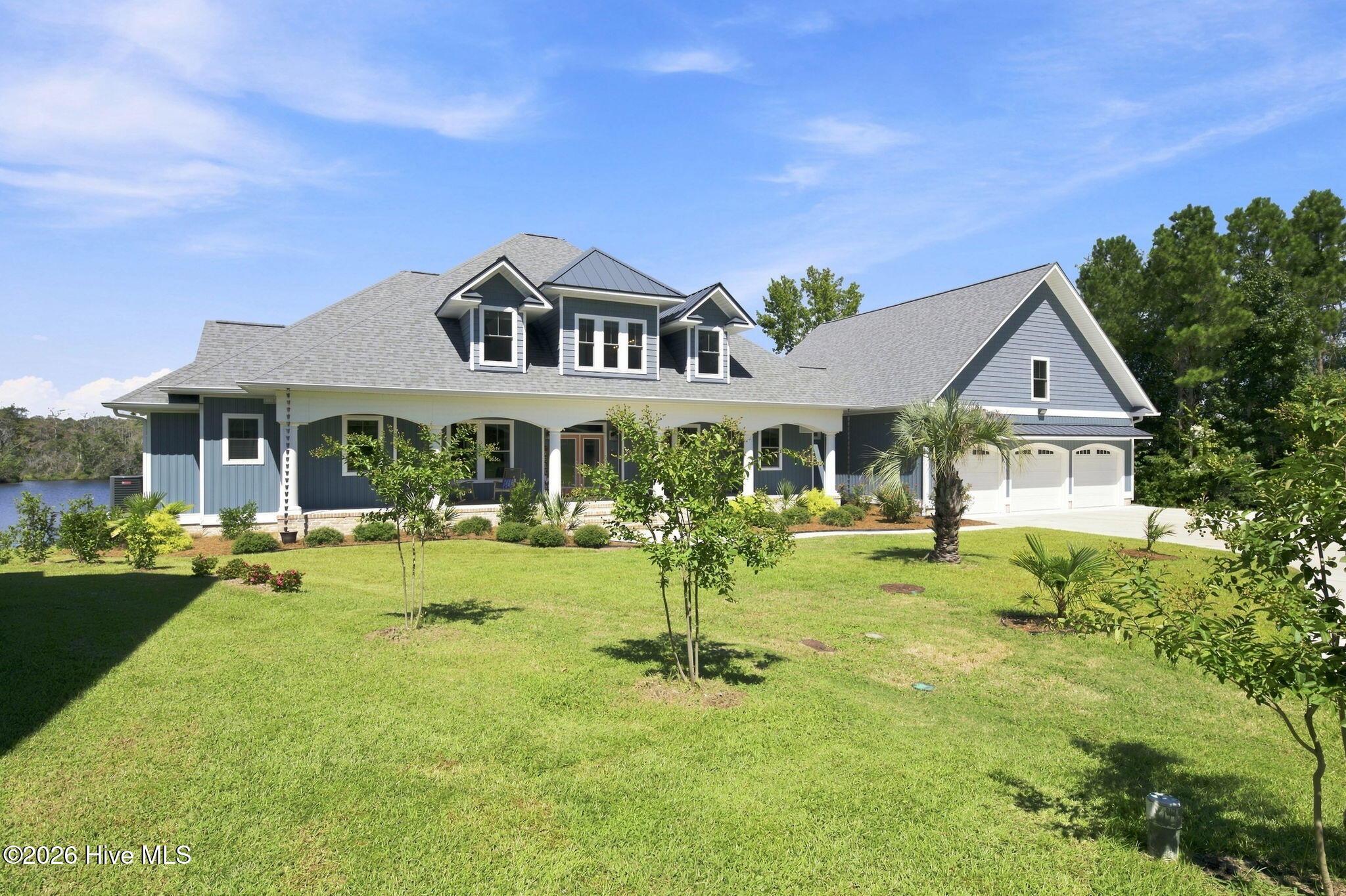 Blue-sided house with a covered porch, three-car garage, landscaped lawn, and water visible beside it.