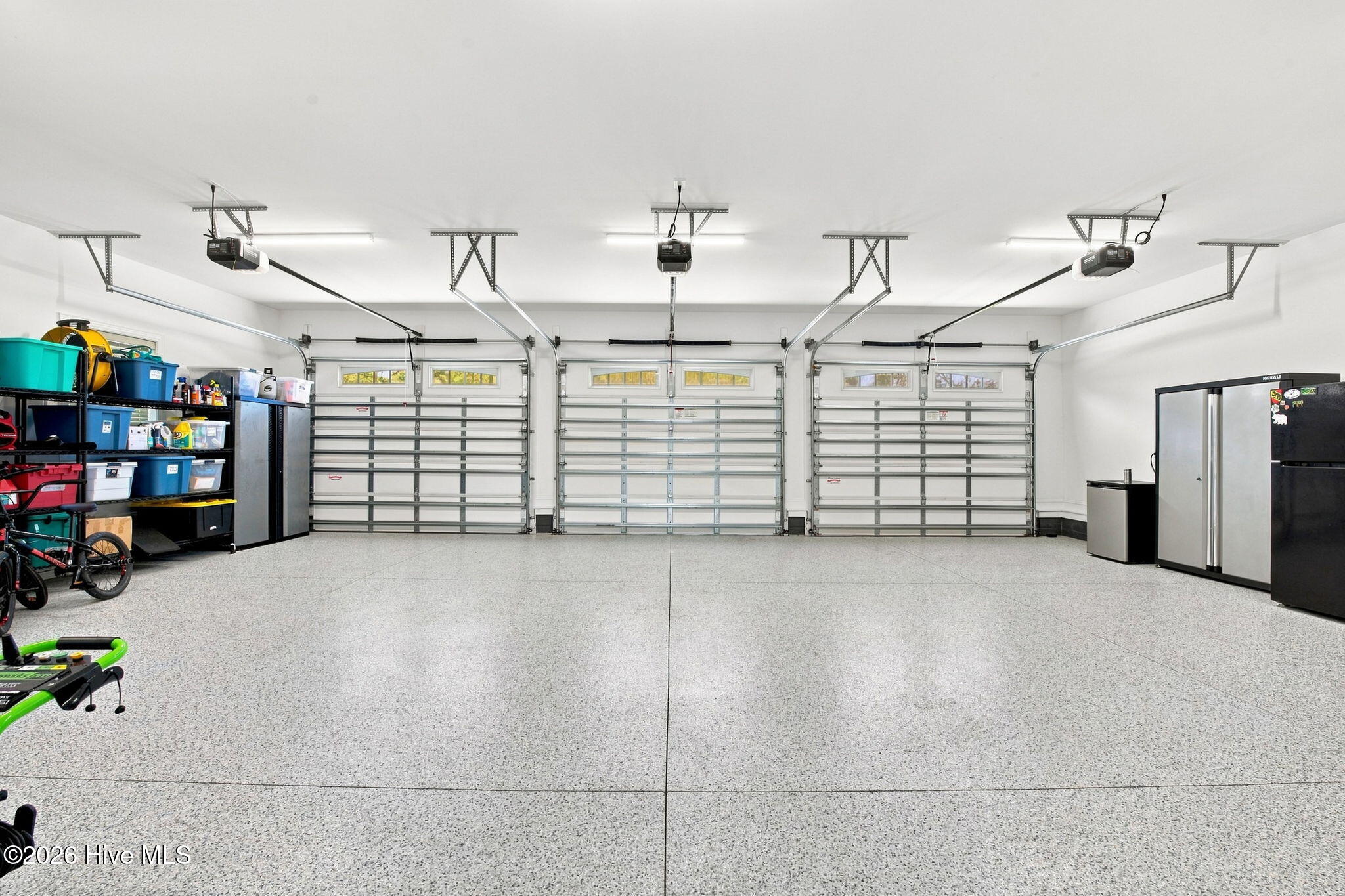 Three-car garage with a speckled floor, storage shelves, and refrigerators.