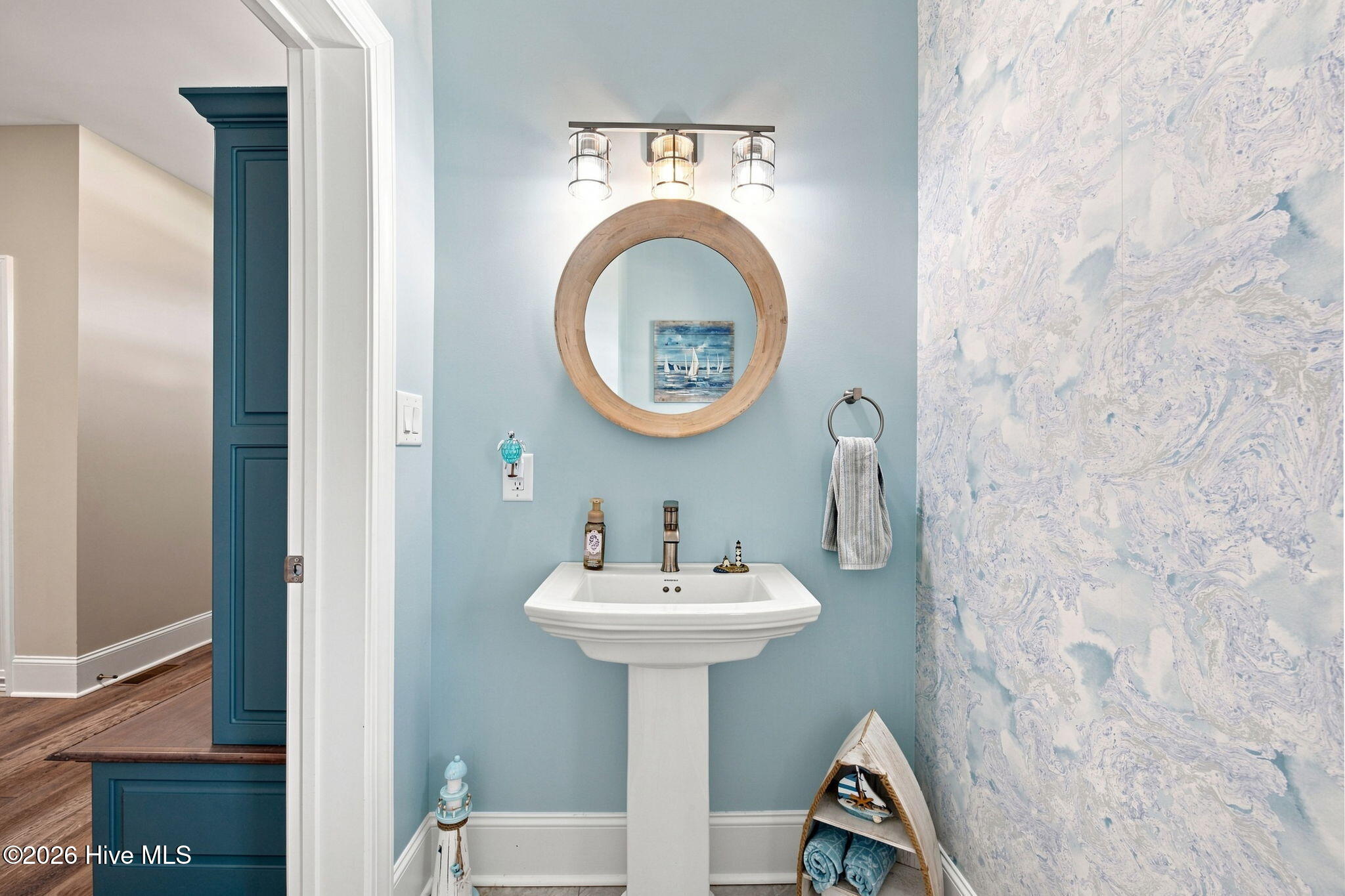 Bathroom with a pedestal sink, round wood-framed mirror, pale blue walls, and patterned wallpaper.