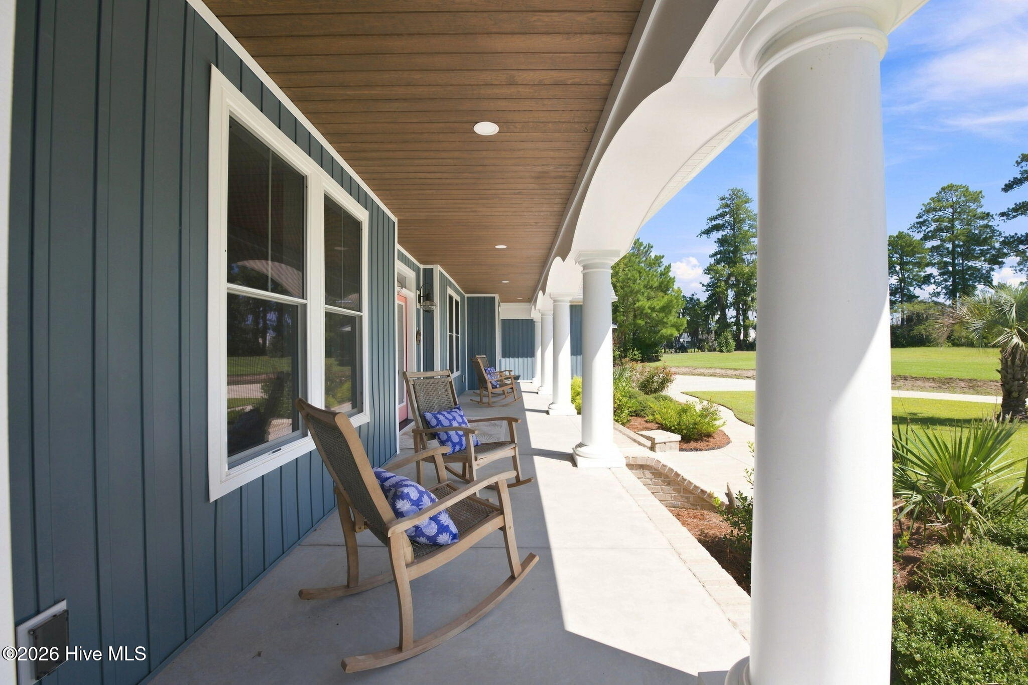 Covered porch with white columns, blue siding, and rocking chairs overlooking a sunny lawn.