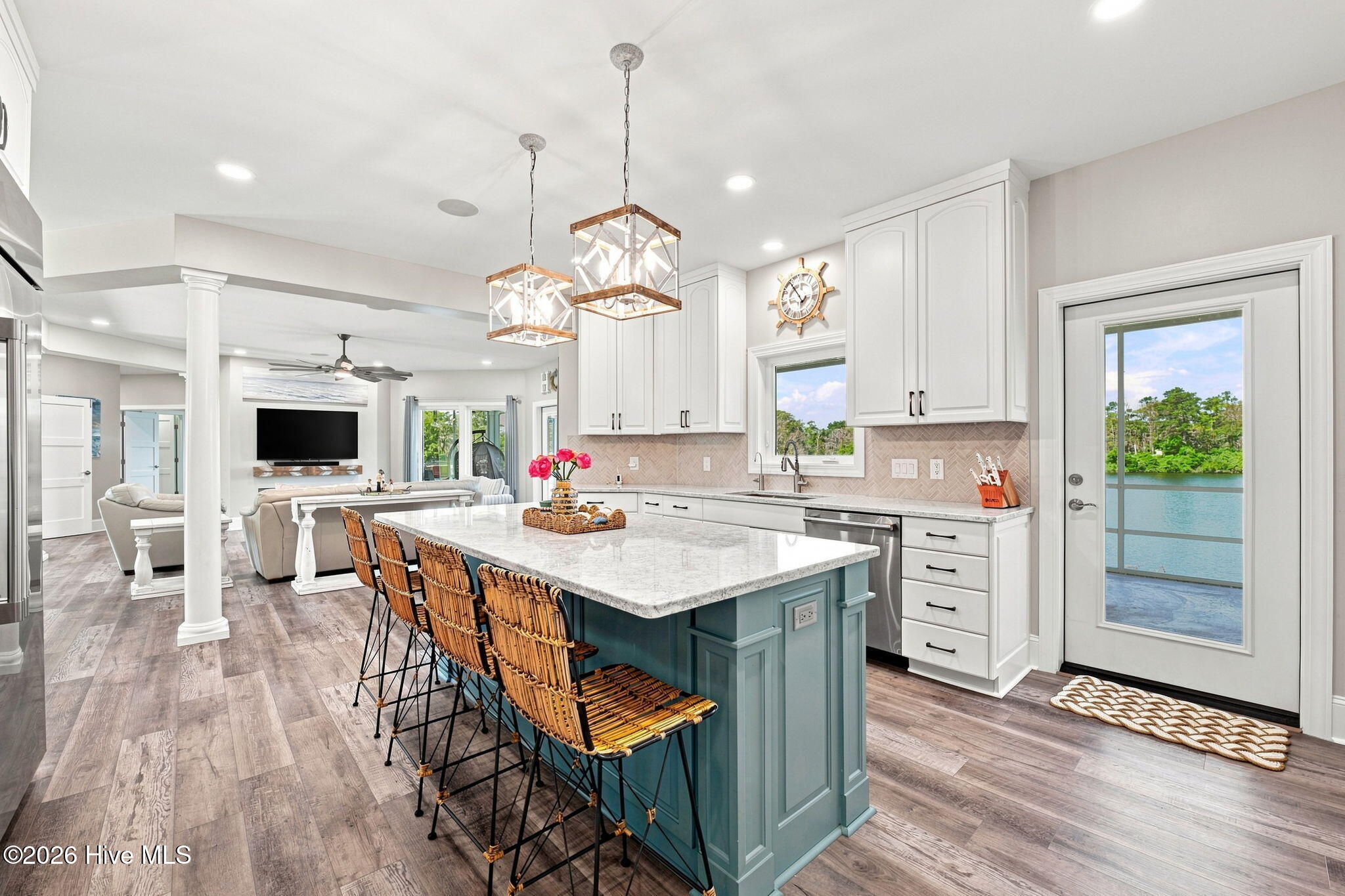 Open-plan kitchen with a large teal island and woven stools, overlooking the water through a glass door.