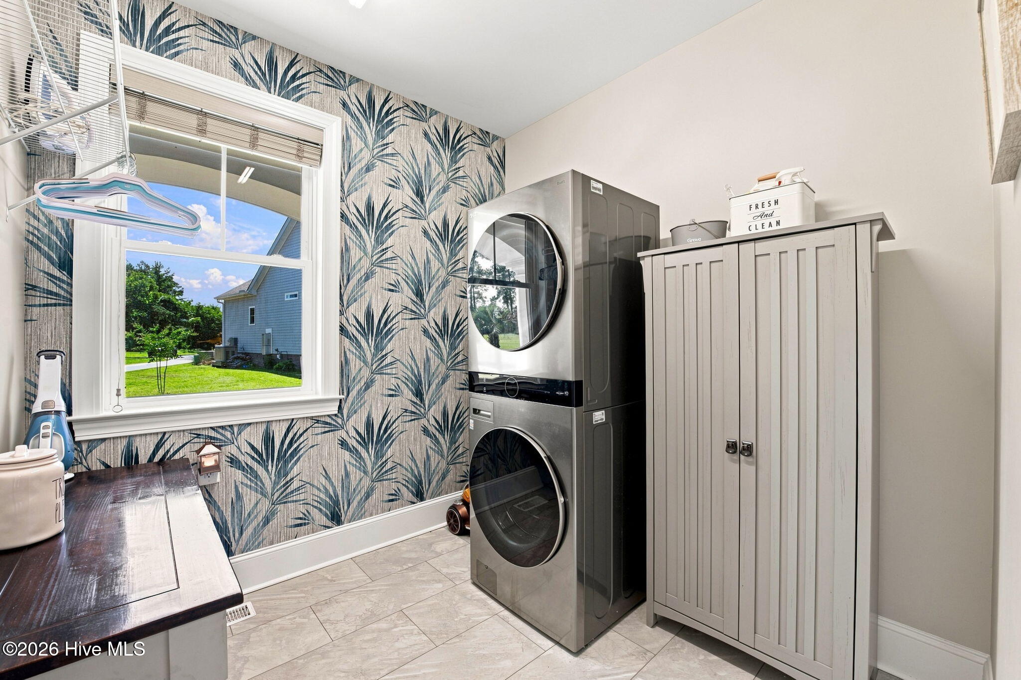 Laundry room with stacked stainless steel washer and dryer beside a gray storage cabinet and palm-leaf wallpaper.