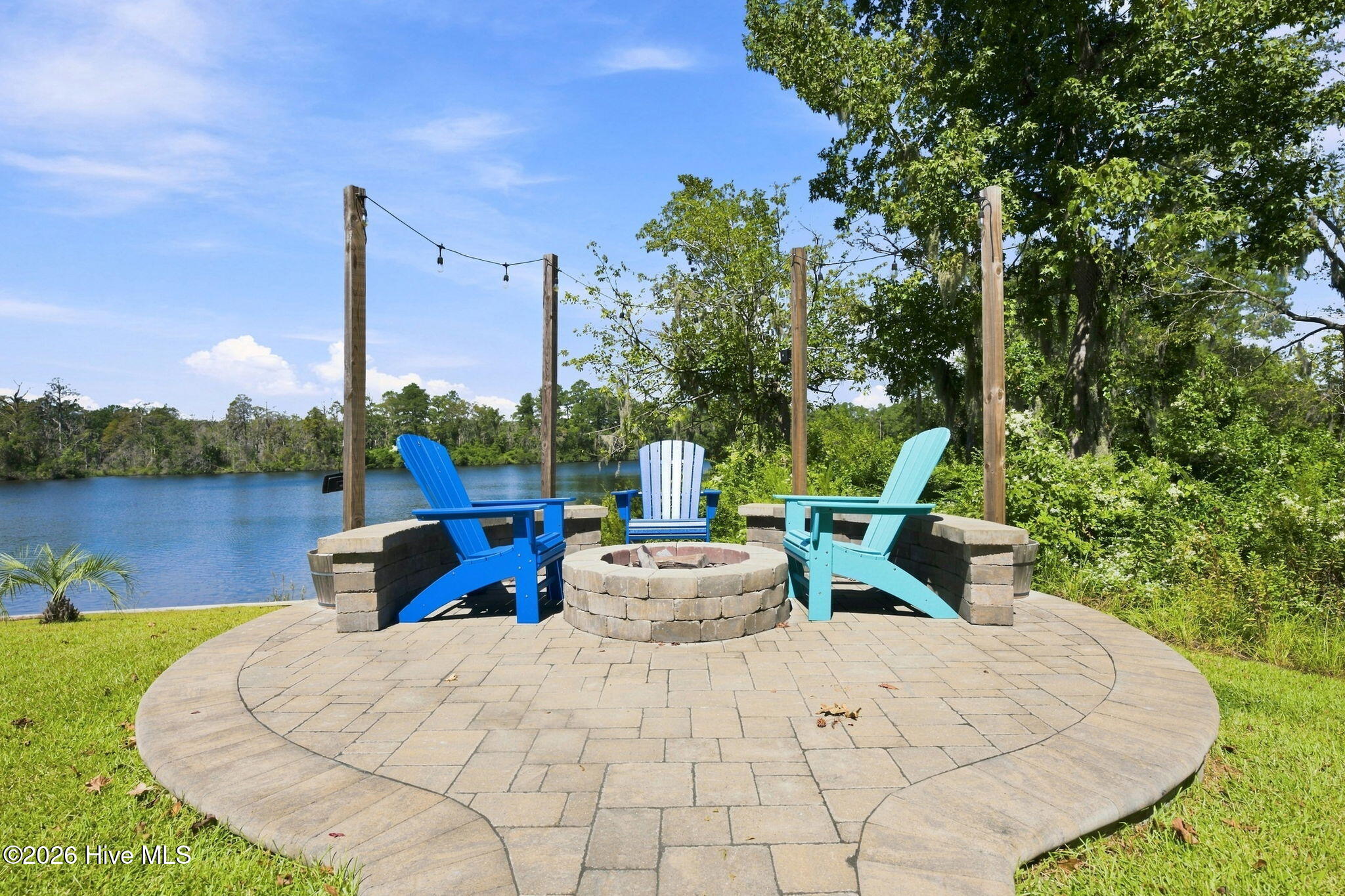 Lakeside patio with a stone fire pit, three colorful Adirondack chairs, and overhead string lights.